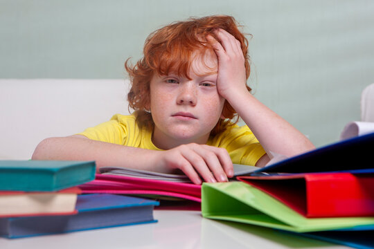 Homework at Home. A young boy looking overwhelmed by his homework. He is holding his head with one hand, a look of boredom on his face, and a large stack of books and folders around him on the table.