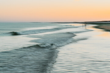 Naklejka premium Long, gentle waves roll onto a quiet beach as the sun sets in the distance.