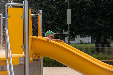 Child in green cap enjoying bright yellow water slide with splashing fountain at modern aqua...