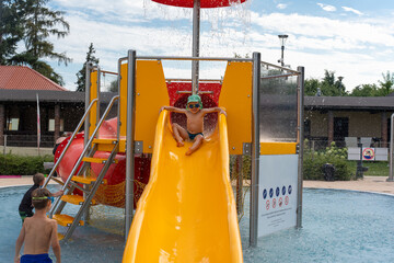 Cheerful child wearing green hat and goggles posing confidently on orange water slide at swimming...