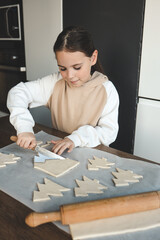 Girl cutting Christmas tree shaped cookies from dough on parchment paper. Child baking holiday...