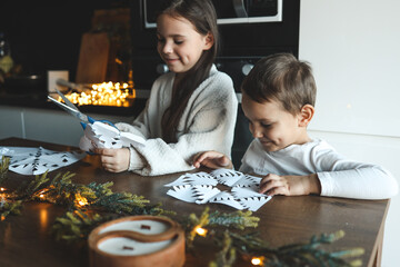 Brother and sister making paper snowflakes at home. Kids cutting and folding white paper, smiling...
