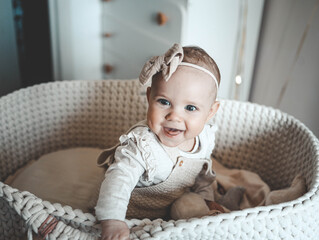 Smiling baby girl sitting in knitted basket crib, wearing beige outfit and bow headband, looking at...