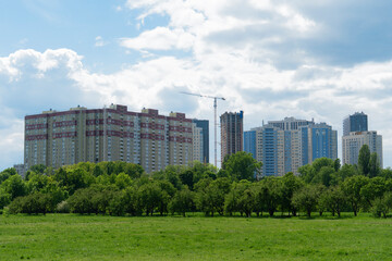 A view of modern multi-story apartment buildings in Kyiv, Ukraine, rising behind lush green trees and a sprawling grassy field under a partly cloudy sky