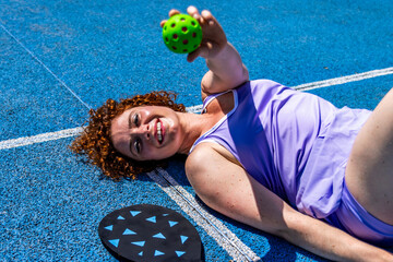 Happy pickleball player lying down on court holding ball and paddle