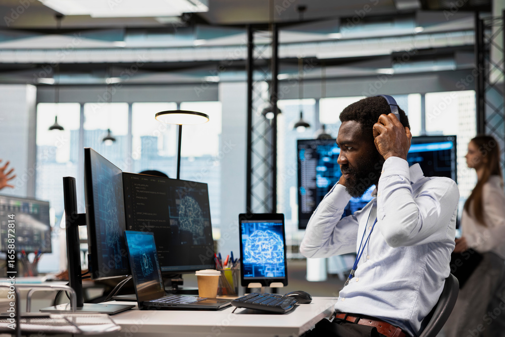 Poster programmer in office relies on ai to run tasks across devices, monitoring progress on screens, liste