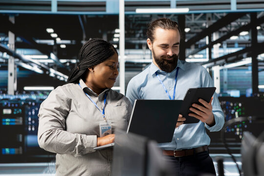 Happy system administrators in data center using devices, verifying firewall configurations. Jolly teamworking IT experts performing equipment vulnerability scans in server hub