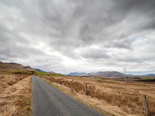 Very narrow asphalt road in a country side with huge wild field with brown grass and mountains in the background, dramatic cloudy sky. Irish nature in Connemara, Ireland.