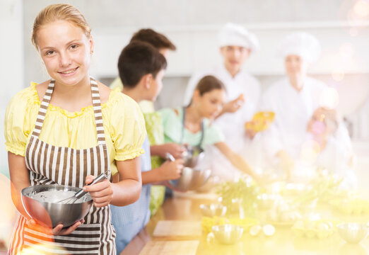 Interested enthusiastic teenage girl participating in culinary class of children group, engrossed in cooking process mirroring professional chef instructor steps