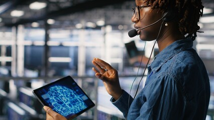 Data center worker on industrial platform speaks in headset microphone while overseeing maintenance on AI rigs below. Admin uses tablet to coordinate deep learning system tasks with team