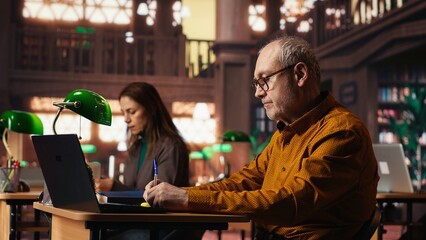 Focused senior person reading web educational papers in a university library, committed to completing his studies and achieving a meaningful degree in his later years. Old man.