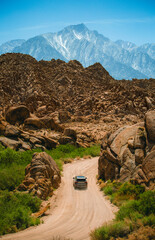 SUV Driving Along Rocky Desert Trail Toward Distant Mountain Peaks Under Clear Blue Sky