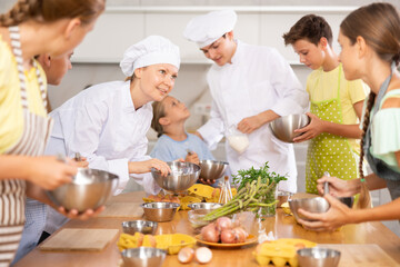 At culinary master class, experienced female chef in white uniform explains to children subtleties...