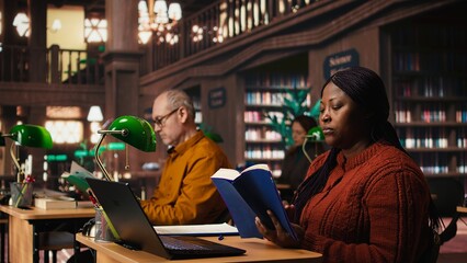 African american woman with a book in hand studying in a quiet library space, working hard to graduate college. Pupil consulting academic literature for master diploma goal.