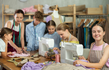 Engaged teenage schoolgirl enjoying group sewing class, stitching at machine in school tailoring studio while peers working with fabric and pattern under guidance of female instructor..