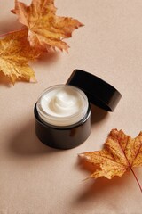 Open black jar of face cream with autumn leaves on neutral background