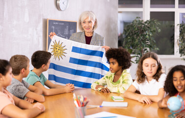 Naklejka na ściany i meble Mature female teacher, conducting a lesson in the classroom, tells the pupils the history of Uruguay and holds the national .flag of the country in her hands