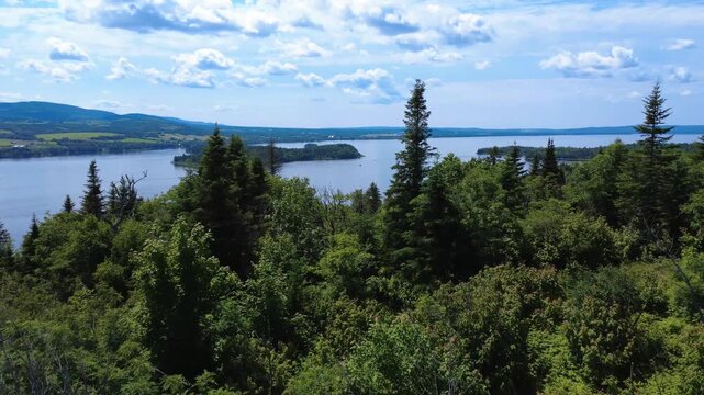 Flight through the treetops of a forest toward an island in a large freshwater lake surrounded by vegetation under a panoramic cloudy sky. Lake Matapedia, Quebec, Canada, 2025.