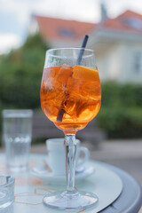 A large glass filled with Aperol Spritz cocktail on a table at an outdoor cafe surrounded by greenery and distant buildings.