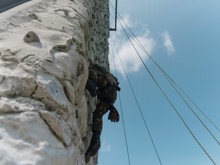 Military personnel rappelling down climbing wall during training exercise with blue sky background