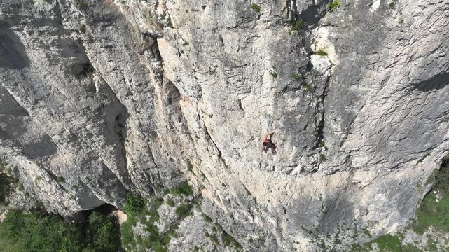 AERIAL: Shirtless man climbs a challenging route in a massive, limestone rock face. Young sport climber appears small against the vertical wall as he tackles a difficult climb on a sunny summer day.