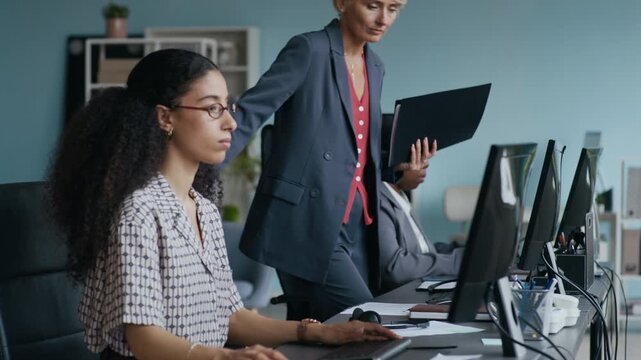 Elegant blond female operator coming up to her workplace, sitting down in armchair between two diverse colleagues, putting headset on her head and looking at computer screen