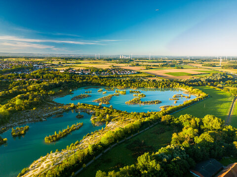 Aerial view of Dyckerhoffsee with turquoise water in Beckum, Germany, contrasting protected nature reserve with industrial Ruhr area skyline of smokestacks and wind turbines on the horizon