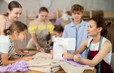 Positive female teacher sitting at worktable in tailoring workshop, demonstrating basics of sewing...
