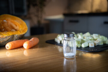 Salt shaker in the foreground with vegetables and kitchen in the background.