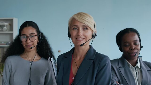 Push in of three young elegant diverse female customer support representatives with headsets looking at camera with smiles while standing in call center