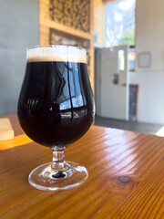 A tall, dark craft beer, a chocolate stout, in a clear pint glass, sitting on a wooden table of a crafted microbrewery. The dark glass of ale has a foamy head on top. The background is wood stacked.