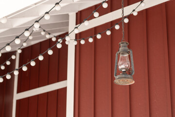 The exterior wall of a red wooden barn with white trim. There are strings of white solar lights hanging from the outside roof of the building. An antique rusty glass hurricane lamp hangs from a chain.