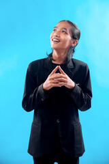 Asian man in a black suit smiles while looking upward with fingers clasped together, expressing...