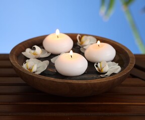 Burning candles and orchid flowers in bowl of water on wooden table against blurred blue...