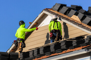 Two roofers prepare roofing materials atop a house under construction, organizing stacks of...