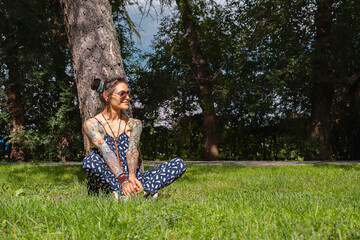 Woman with tattoos and sunglasses sitting relaxed by tree in sunlit park with green grass.