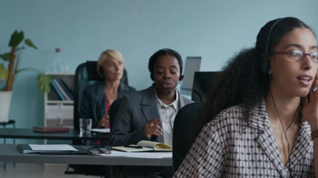 Row of young intercultural female customer support representatives sitting in coworking space and consulting online clients while taking notes in notepads
