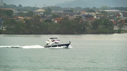 Obraz premium Luxury motor yacht speeds across the water near a coastal town on a bright day, leaving a white wake