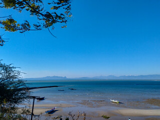 Scenic tropical beach with calm turquoise sea, fishing boats on sandy shore, and distant mountain range under a bright clear blue sky
