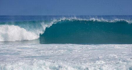 Dramatic turquoise wave crashing onto a sandy beach under a
clear blue sky, with foamy water and dynamic sea spray.