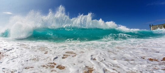 Dramatic turquoise wave crashing onto a sandy beach under a clear blue sky, with foamy water and dynamic sea spray.
