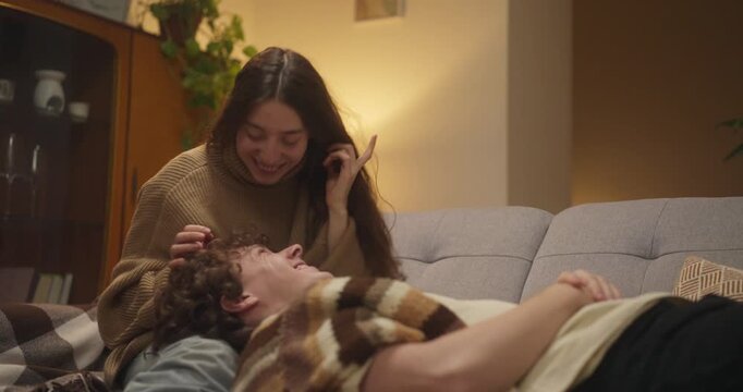 Girl in sweater kisses her boyfriend on the forehead when he lies on her lap during cozy chat and romantic evening on cozy sofa at home in the evening