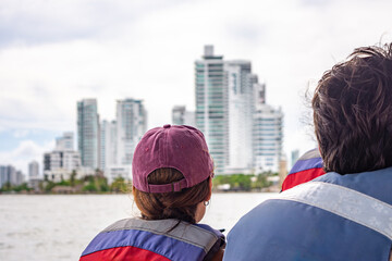 A couple wearing life vests enjoys a boat ride with modern buildings of Cartagena, Colombia, in the...