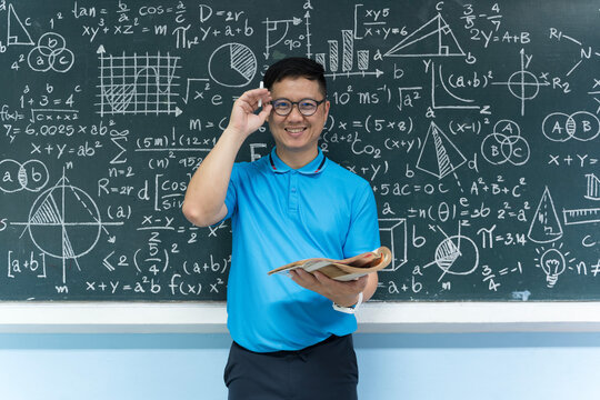 Smiling mathematics teacher holding book in front of blackboard full of formulas and equations in class