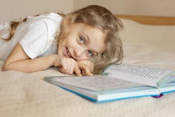 A cheerful young girl is lying on a cozy bed, smiling enthusiastically while reading a book....