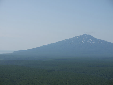 Snowcapped Bogdan Hmelnicky volcano with snow in Iturup green summer landscape with atmospheric haze on sunny day, Kuril islands