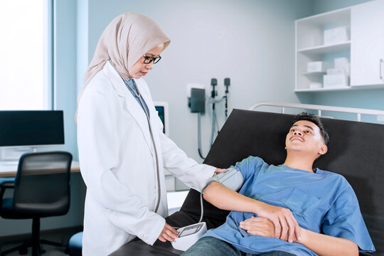 Female doctor measuring a patient's blood pressure. A female doctor wearing a headscarf, stethoscope, and lab coat is using a blood pressure monitor on a male patient lying on a hospital bed.