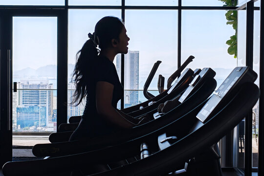 Silhouette of a woman exercising on a treadmill. A silhouette of a woman is exercising on a treadmill in a gym with large windows in the background showing a bright cityscape.