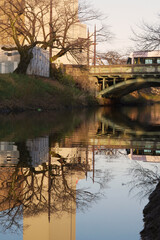 Obraz premium Autumn reflection on a calm river. A bridge, bare trees, and a building are reflected in the smooth surface of the water on a quiet autumn day