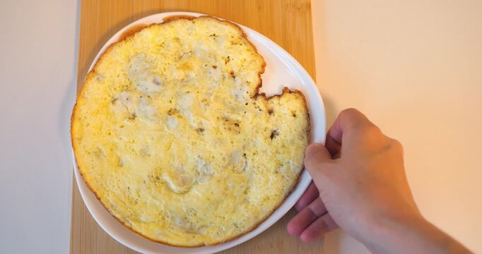 Hand adjusting large omelette on white plate over wooden cutting board, closeup overhead shot in warm kitchen light, breakfast preparation moment.